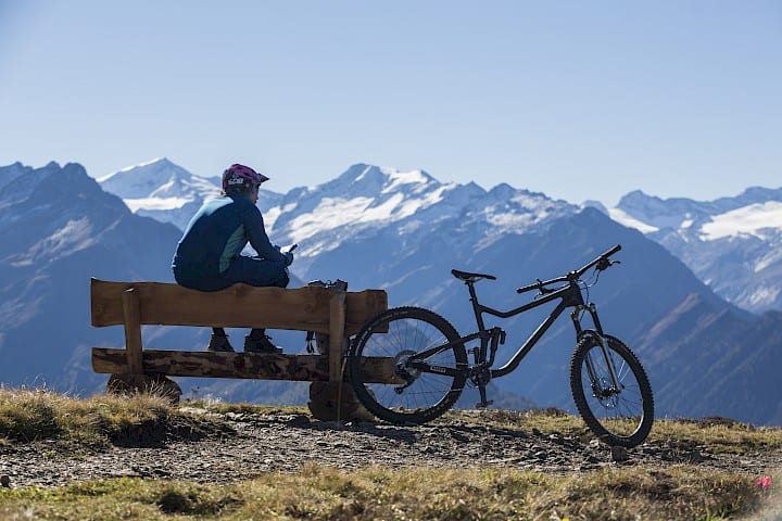 Horský cyklista odpočívá na panoramatické lavičce s výhledem na zasněžené vrcholky Vysokých Taur – pohodová e-bike nebo MTB túra ve Wildkogel Areně s dechberoucím výhledem.
