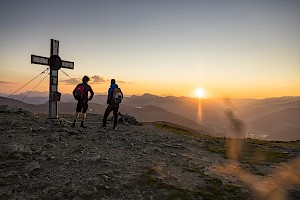 Zwei Wanderer stehen am Gipfelkreuz in der Wildkogel-Arena und blicken in der Morgendämmerung auf das weite Tal und die umliegenden Berge.