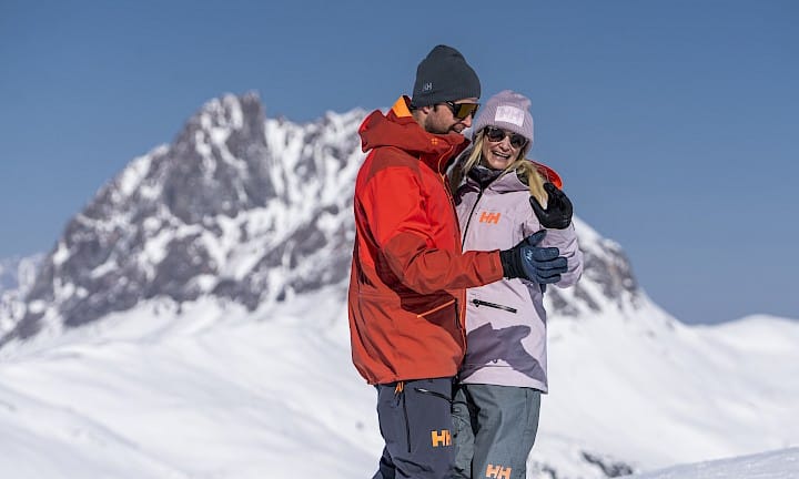 Usměvavý pár tančí Schneewalzer (sněhový valčík) na slunci před velkolepou zimní horskou scenérií. V teplém lyžařském oblečení si užívají romantickou chvíli – ideální pro balíček „Schneewalzer“.