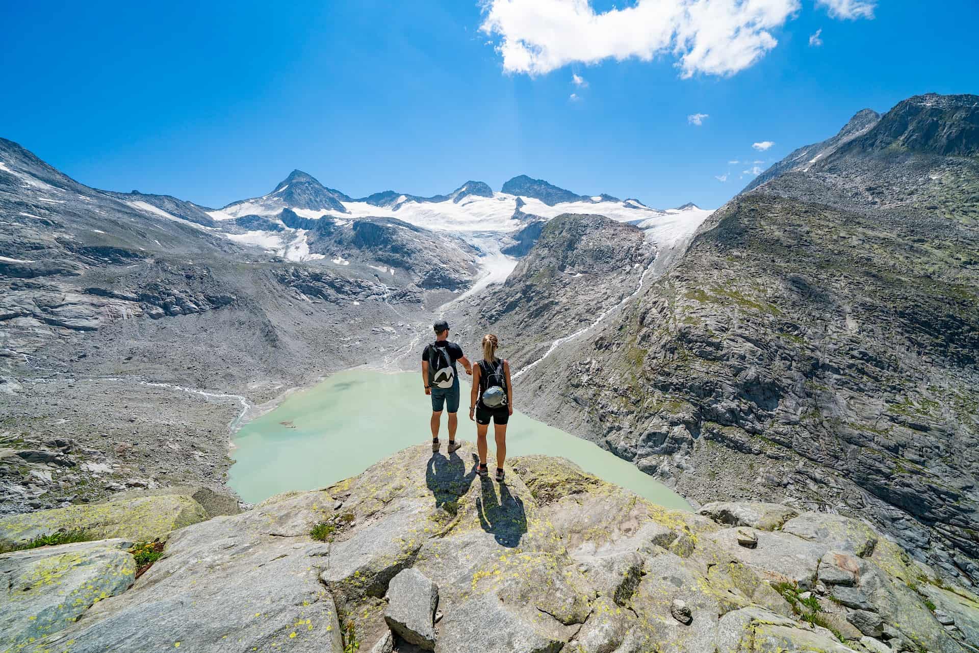 Dva turisté si užívají panoramatický výhled na tyrkysové ledovcové jezero a zasněžené vrcholy v Národním parku Vysoké Taury – přírodní zážitek a dobrodružství v rakouských Alpách.