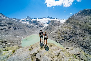 Zwei Wanderer genießen das Panorama über den türkisgrünen Gletschersee und das vergletscherte Hochgebirge im Nationalpark Hohe Tauern – Naturerlebnis und Abenteuer in den österreichischen Alpen.