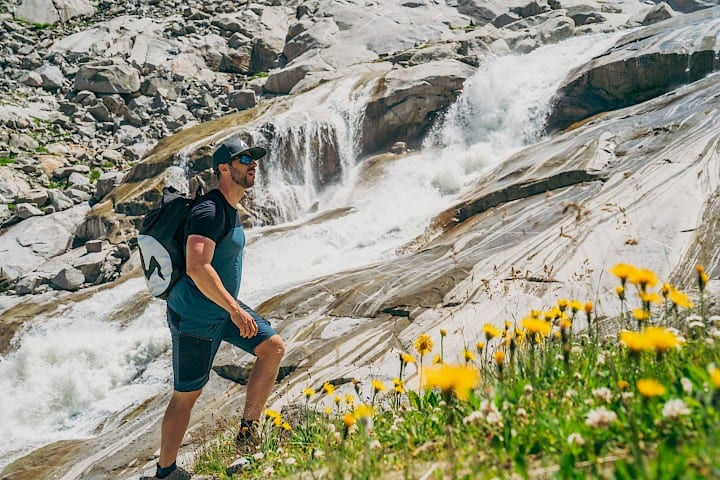 Wanderer an einem Wasserfall im Nationalpark Hohe Tauern – imposante Felsformationen, tosende Gischt und blühende Almwiesen.