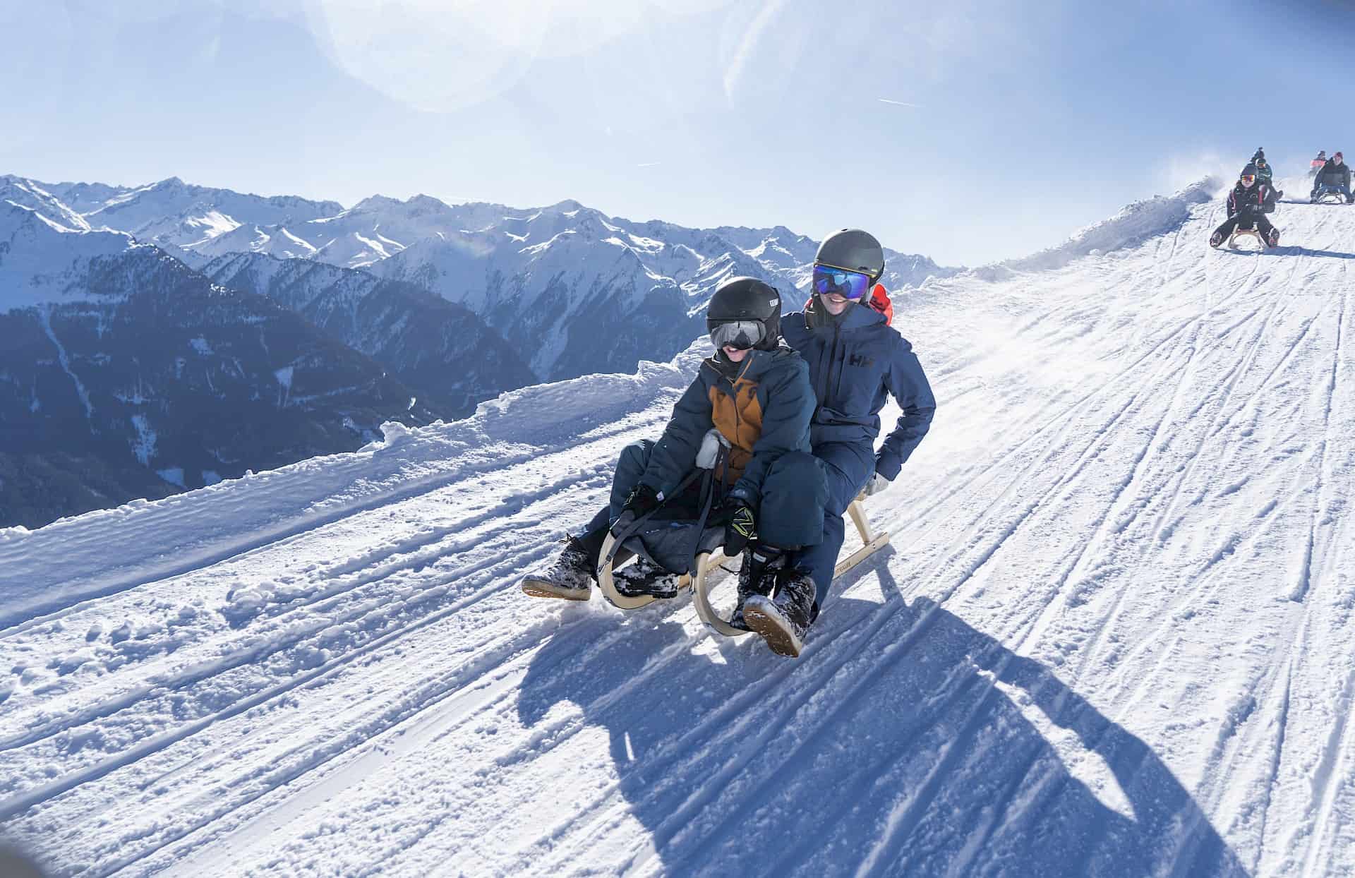 Rodelspaß auf der längsten beleuchteten Rodelbahn der Welt in der Wildkogel-Arena in Bramberg – zwei Personen auf dem Schlitten genießen die verschneite Winterlandschaft und sonniges Wetter in den Hohen Tauern.