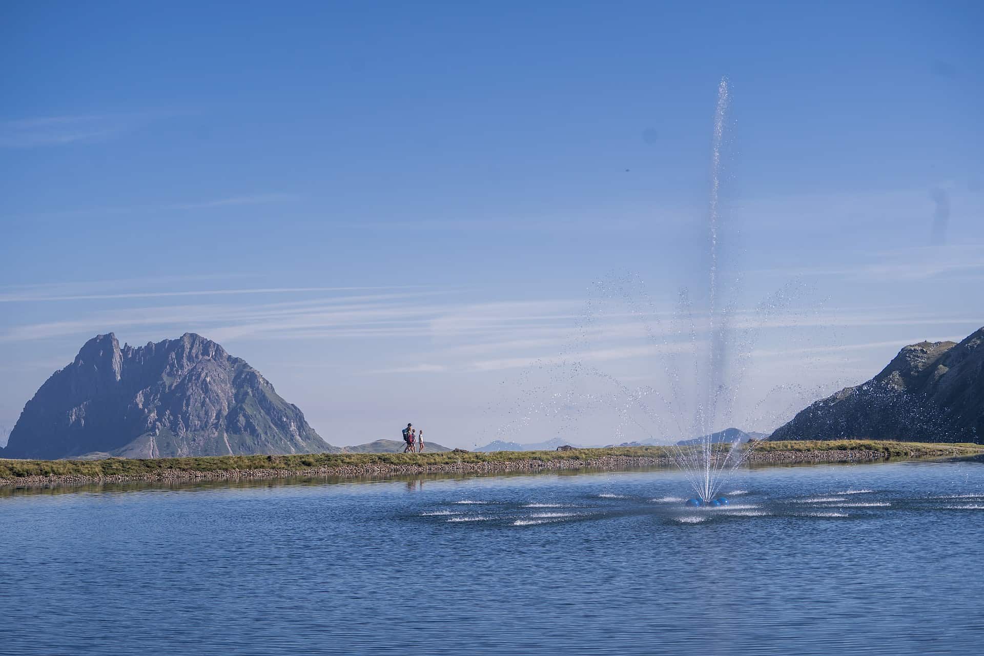 Ein Wanderpaar spaziert entlang eines idyllischen Bergsees mit Wasserfontäne, im Hintergrund ragt der markante Gipfel des Großen Rettensteins in den Himmel – ein entspannter Sommertag im Höhenwandergebiet Wildkogel inmitten der Kitzbüheler Alpen.