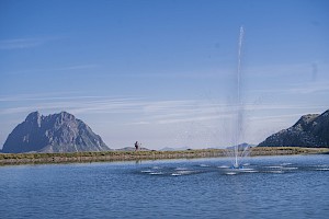 Ein Wanderpaar spaziert entlang eines idyllischen Bergsees mit Wasserfontäne, im Hintergrund ragt der markante Gipfel des Großen Rettensteins in den Himmel – ein entspannter Sommertag im Höhenwandergebiet Wildkogel inmitten der Kitzbüheler Alpen.