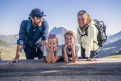 Veselá rodina se dvěma dětmi si užívá přestávku při túře ve vysokohorském turistickém regionu Wildkogel. Všichni čtyři se smějí a opírají o dřevěnou lavičku, v pozadí výrazný vrchol Großer Rettenstein pod modrou oblohou – dokonalý rodinný čas v přírodě.