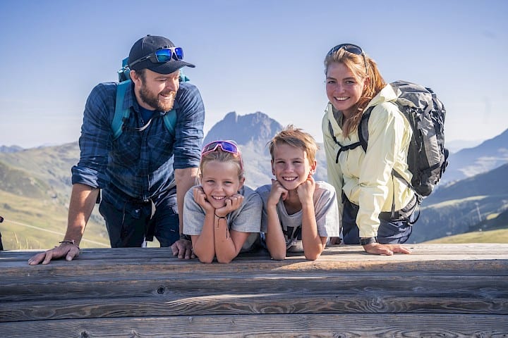 Veselá rodina se dvěma dětmi si užívá přestávku při túře ve vysokohorském turistickém regionu Wildkogel. Všichni čtyři se smějí a opírají o dřevěnou lavičku, v pozadí výrazný vrchol Großer Rettenstein pod modrou oblohou – dokonalý rodinný čas v přírodě.