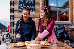 Gemütliche Hüttenpause im Habachtal: Zwei Frauen genießen regionale Schmankerl wie Kaiserschmarrn und eine Brettljause auf der Sonnenterrasse mit Alpenblick.