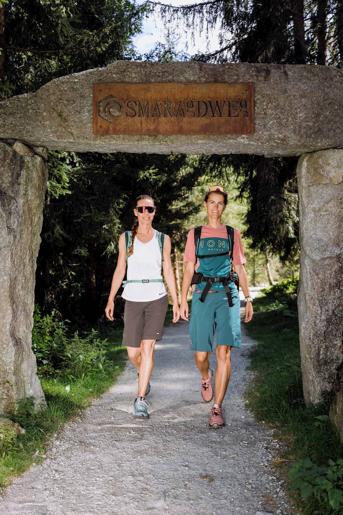 Zwei Wanderinnen gehen durch den steinernen Torbogen mit der Aufschrift „Smaragdweg“ im Habachtal, umgeben von Wald und Sonnenlicht.