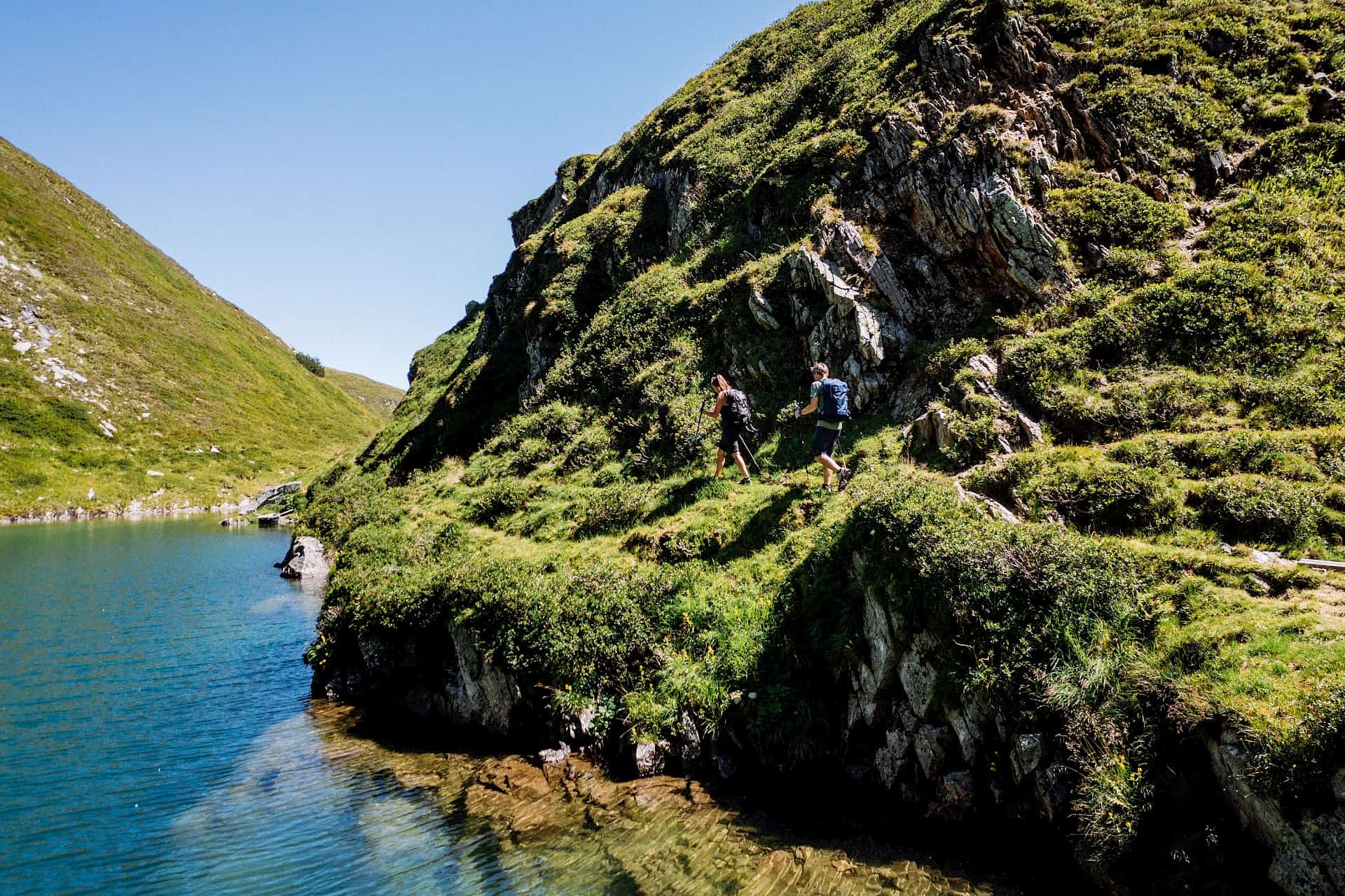 Zwei Wanderer auf einem schmalen Pfad am türkisgrünen Seebachsee im Nationalpark Hohe Tauern, umgeben von schroffen Felsen und grünen Almwiesen – ein echtes Naturerlebnis beim Wandern in der Wildkogel-Arena.