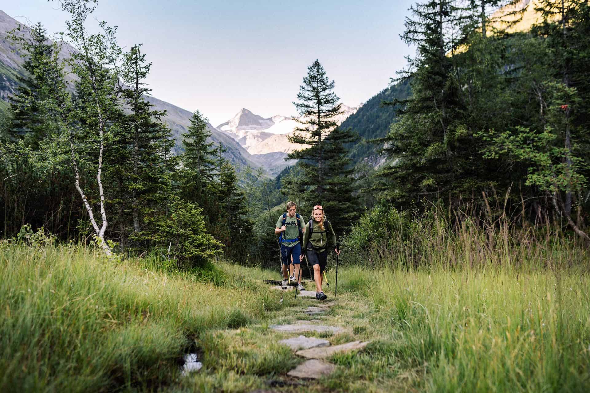 Dva turisté na přírodní horské stezce zeleným vysokohorským údolím Vysokých Taur – letní túra v nedotčené alpské krajině s výhledem na ledovce.